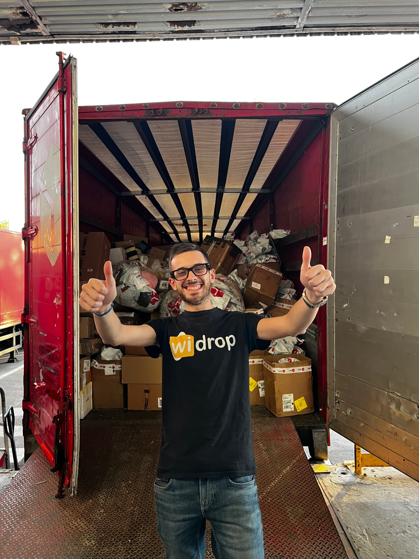 Ragazzo del team Widrop con doppio pollice in su davanti a un tir carico di pacchi al molo di carico